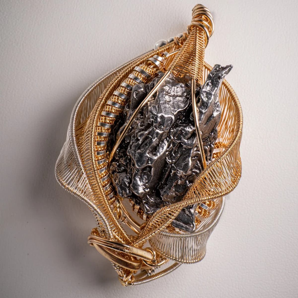 Gjennomsiktig amulett i viklet gulltråd med mørke metallfragmenter fra en meteoritt i midten, sett ovenfra mot lys bakgrunn