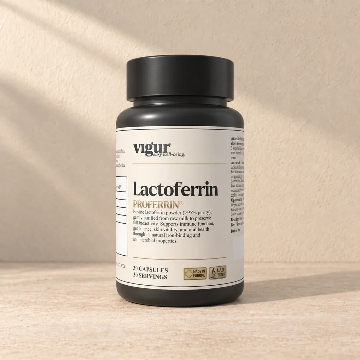 Svart tilskuddsboks med lys etikett merket Vigur og «Lactoferrin PROFERRIN» på beige bakgrunn